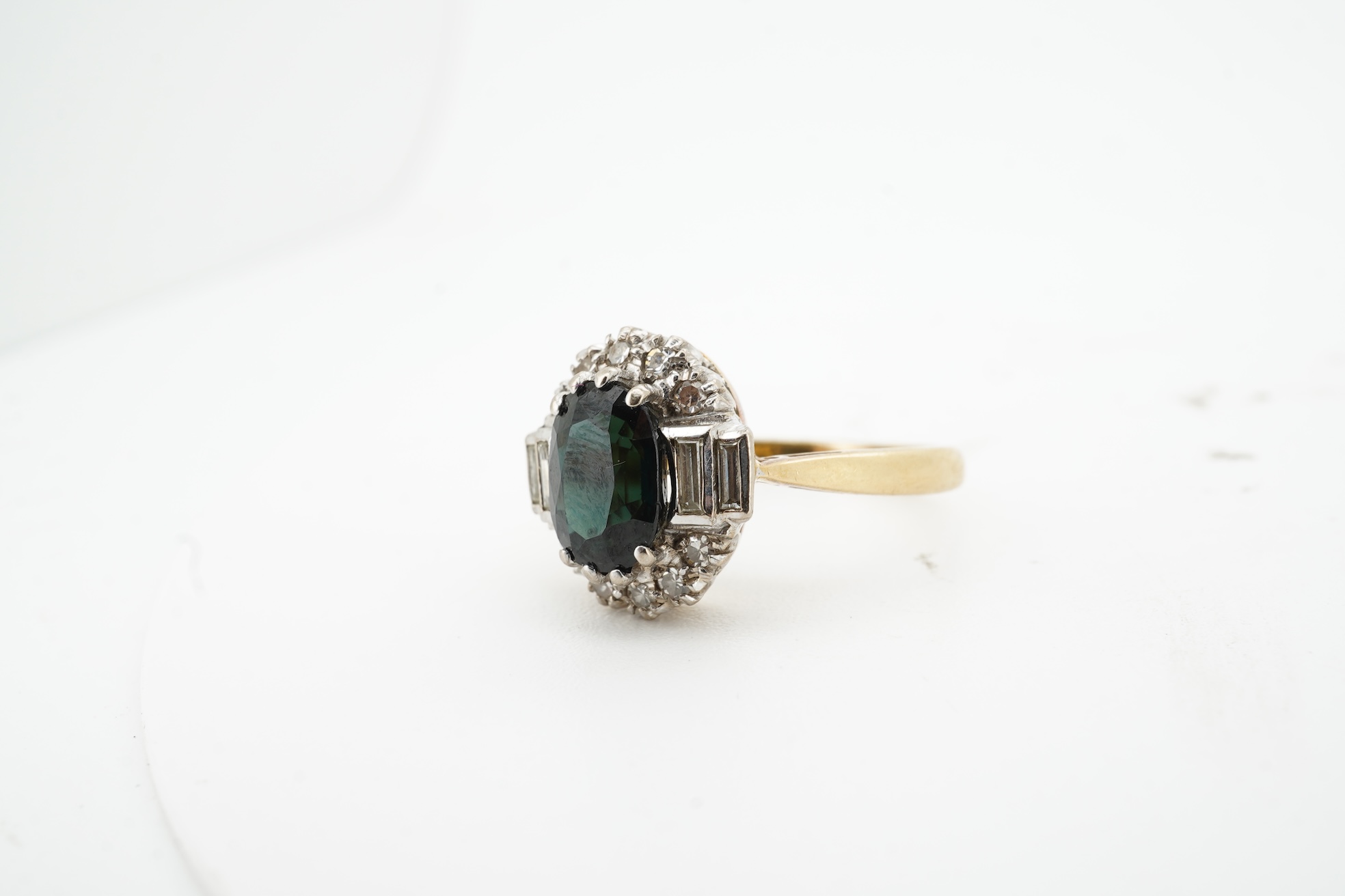 A sapphire and diamond cluster ring, claw-set with an oval sapphire measuring approximately 8.2 x 6.1 x 4.3mm, within a border of brilliant-cut and baguette diamonds totalling approximately 0.20 carats, mounted in 18ct g